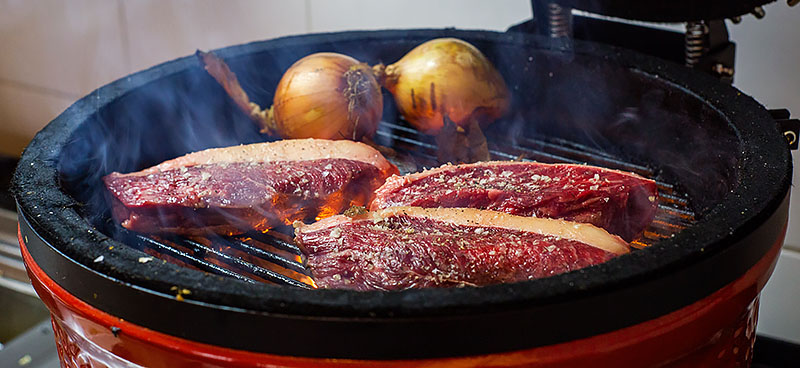 Man sieht einen MONOLITH Keramikgrill, der mit frischem Fleisch und ganzen Zwiebeln bestückt ist. Stichwort direktes Grillen.